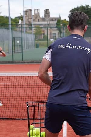 A coach assisting a young girl during a tennis practice session on a red clay court at The Lensbury, with a historic building in the background.