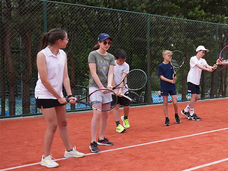 Children holding tennis rackets during a junior tennis camp at The Lensbury, standing on a clay court with a green fence in the background.
