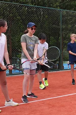 Children holding tennis rackets during a junior tennis camp at The Lensbury, standing on a clay court with a green fence in the background.