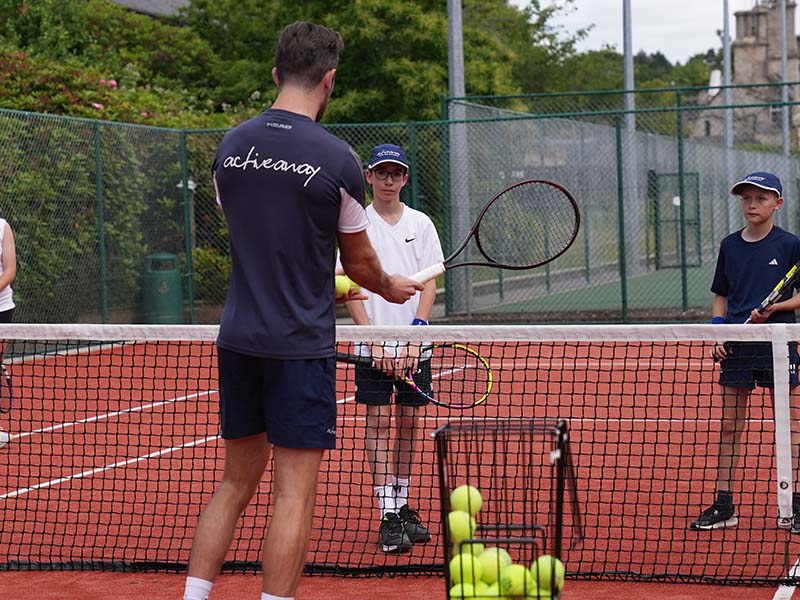 Tennis coach teaching juniors at The Lensbury tennis camp, with players holding rackets on a red clay court.
