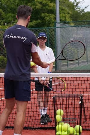 Tennis coach teaching juniors at The Lensbury tennis camp, with players holding rackets on a red clay court.