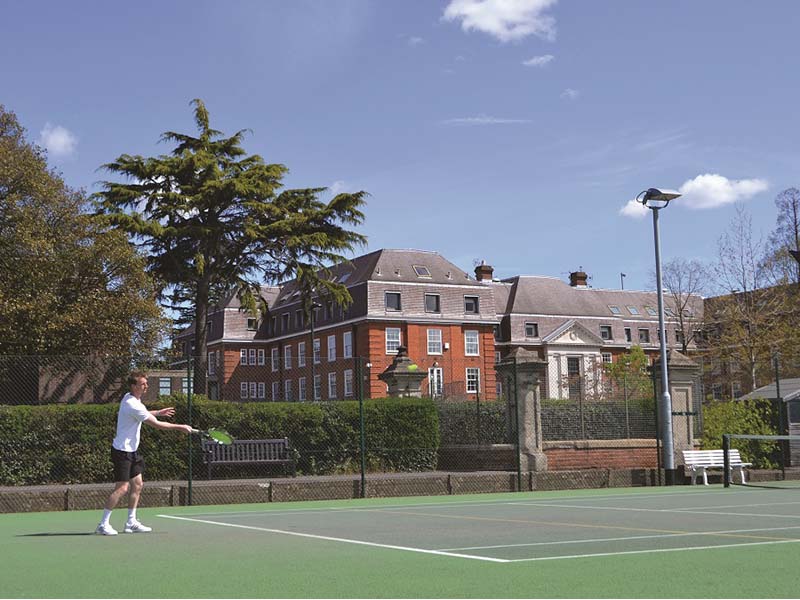 A young player poised to hit a forehand on an outdoor tennis court at The Lensbury, with a red-brick building and greenery in the background.