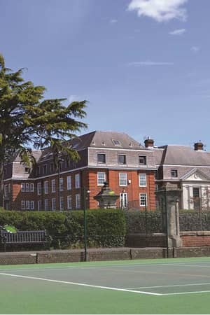 A young player poised to hit a forehand on an outdoor tennis court at The Lensbury, with a red-brick building and greenery in the background.