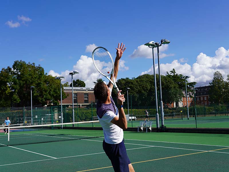 Young tennis player serving on an outdoor court at The Lensbury, with trees and buildings in the background on a bright sunny day.