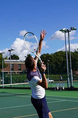 Young tennis player serving on an outdoor court at The Lensbury, with trees and buildings in the background on a bright sunny day.