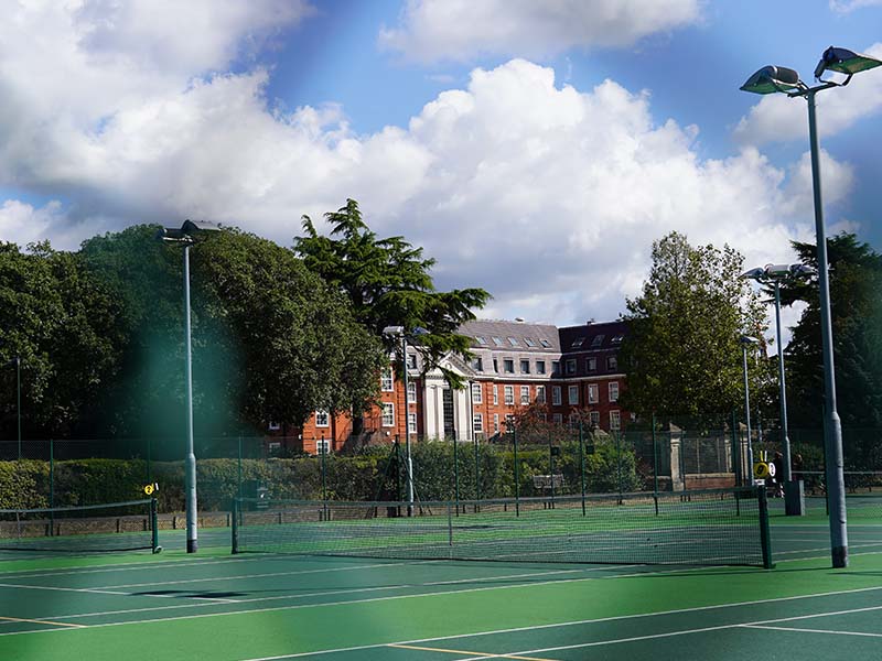 Tennis court at The Lensbury Club with green surface, surrounded by trees and the clubhouse in the background.