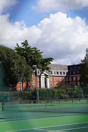 Tennis court at The Lensbury Club with green surface, surrounded by trees and the clubhouse in the background.