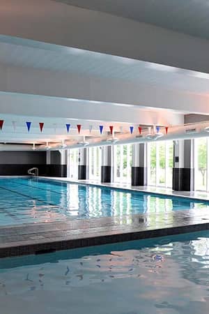 Indoor swimming pool at The Lensbury with large windows, black and white tiling, and colourful triangular flags overhead.