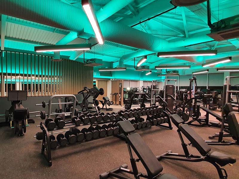 Interior view of The Lensbury gym with free weights, benches, and exercise machines under teal lighting.