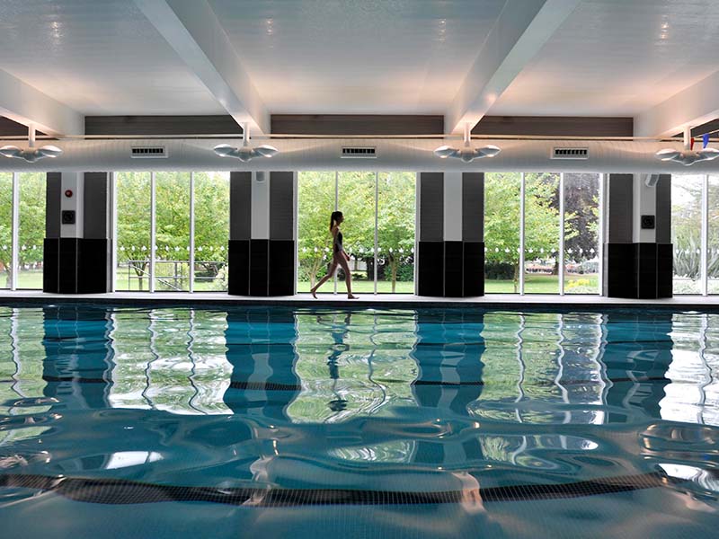 Child walking by an indoor swimming pool with large windows and views of greenery at The Lensbury sports club.