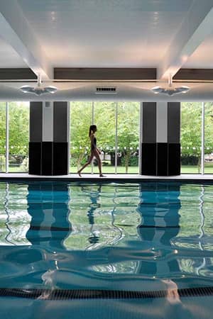 Child walking by an indoor swimming pool with large windows and views of greenery at The Lensbury sports club.