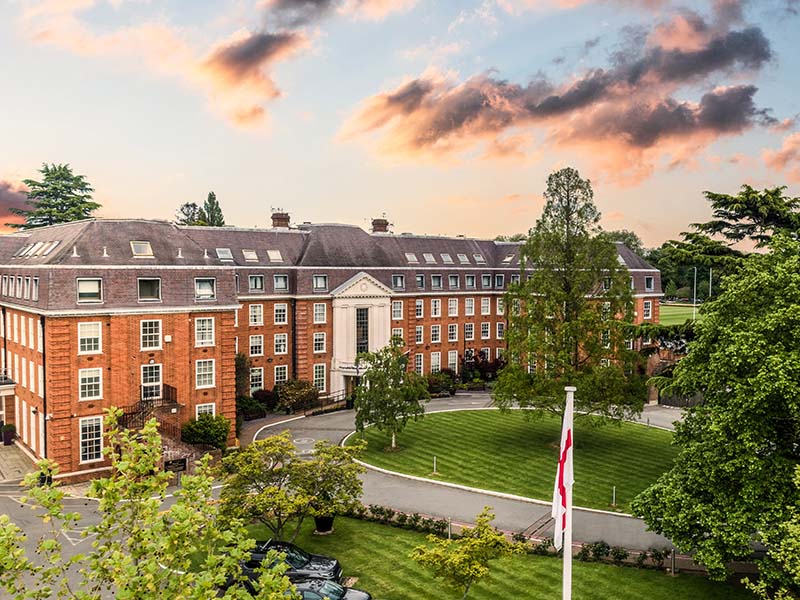 The Lensbury Club in Teddington, UK, a luxurious red-brick building with manicured lawns and trees during sunset.