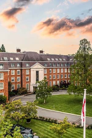 The Lensbury Club in Teddington, UK, a luxurious red-brick building with manicured lawns and trees during sunset.