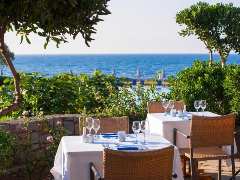 Outdoor dining area at Kalimera Kriti with tables set for dining, overlooking the sea through lush greenery.