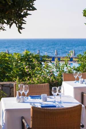 Outdoor dining area at Kalimera Kriti with tables set for dining, overlooking the sea through lush greenery.