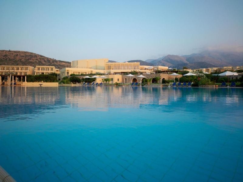 View of the Kalimera Kriti Hotel's swimming pool with sun loungers, parasols, and mountains in the background.