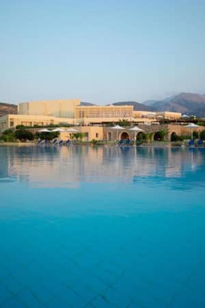 View of the Kalimera Kriti Hotel's swimming pool with sun loungers, parasols, and mountains in the background.