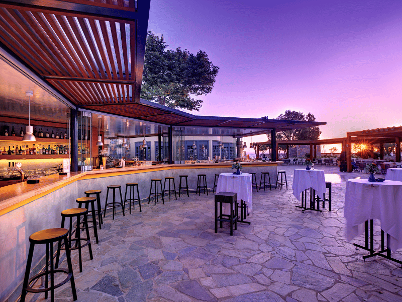 Outdoor bar area at Kalimera Kriti Resort with wooden stools, stone-tiled floor, and a sunset view.