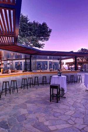 Outdoor bar area at Kalimera Kriti Resort with wooden stools, stone-tiled floor, and a sunset view.