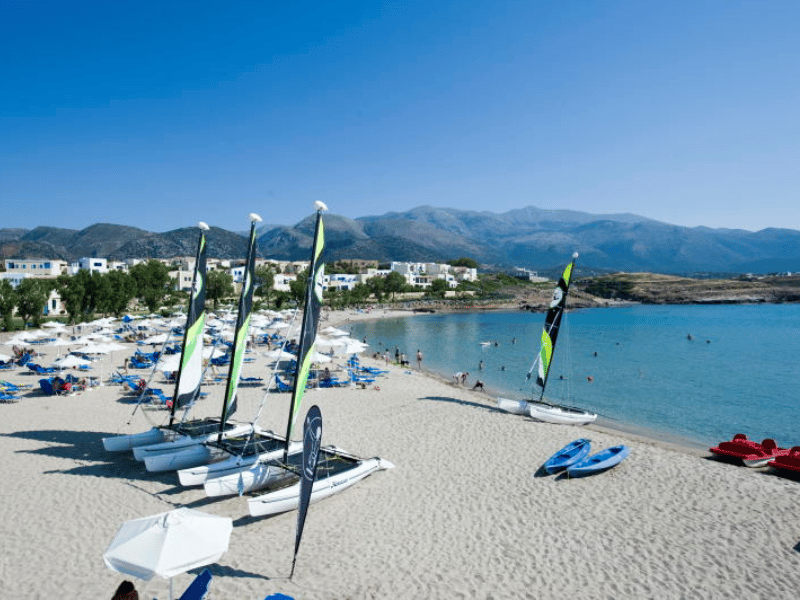 View of Kalimera Kriti beach in Crete, with catamarans, sun loungers, parasols, and a mountainous backdrop under a clear blue sky.