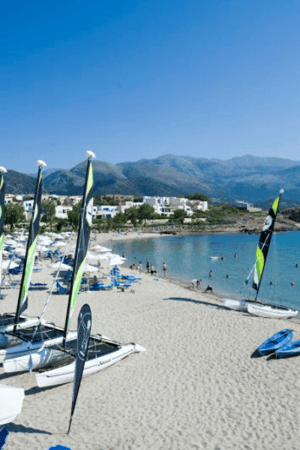 View of Kalimera Kriti beach in Crete, with catamarans, sun loungers, parasols, and a mountainous backdrop under a clear blue sky.