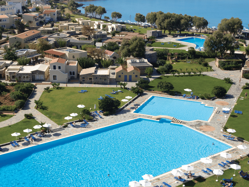 Aerial shot of Kalimera Kriti Resort's swimming pool with sea view, surrounded by greenery and traditional architecture.