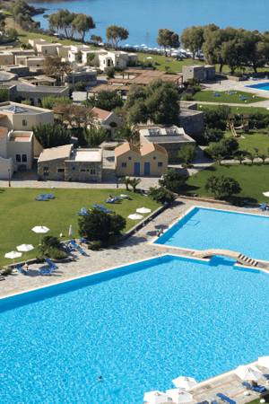 Aerial shot of Kalimera Kriti Resort's swimming pool with sea view, surrounded by greenery and traditional architecture.