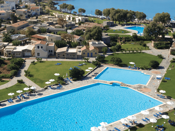 Aerial shot of Kalimera Kriti Resort's swimming pool with sea view, surrounded by greenery and traditional architecture.