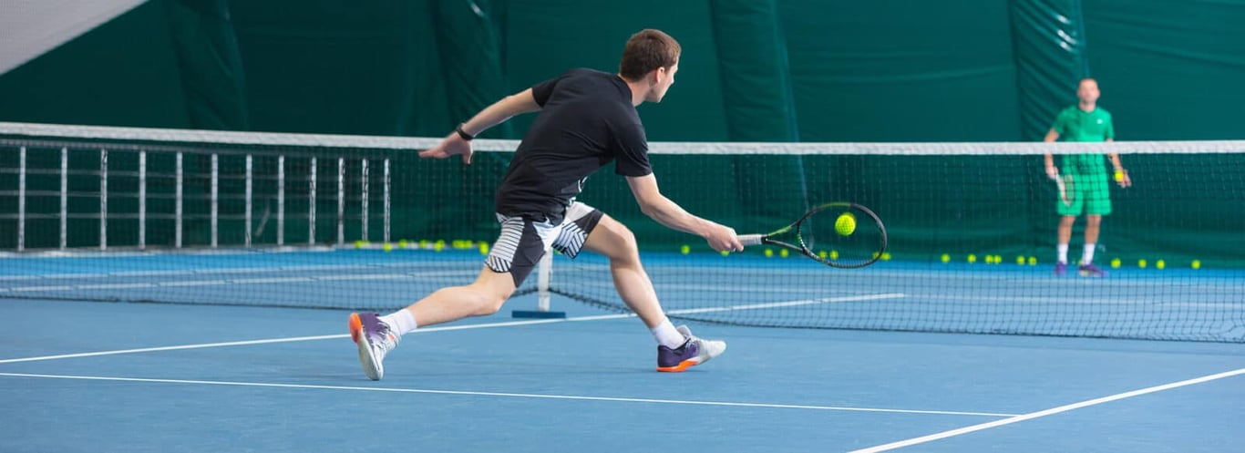 Young man playing tennis indoors, reaching for a backhand shot during a game, with an opponent waiting on the other side of the net.