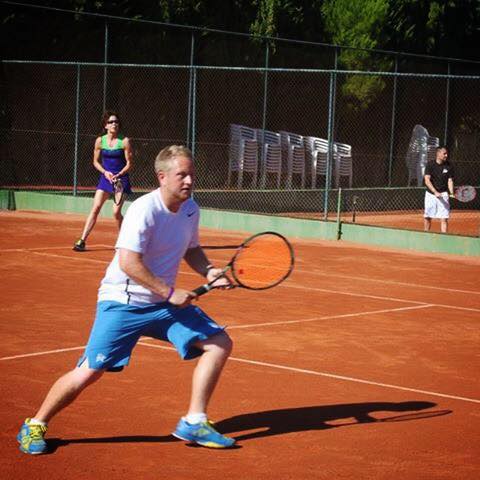 Kieran Loveder preparing for a forehand shot on a clay tennis court, with two other players in the background.