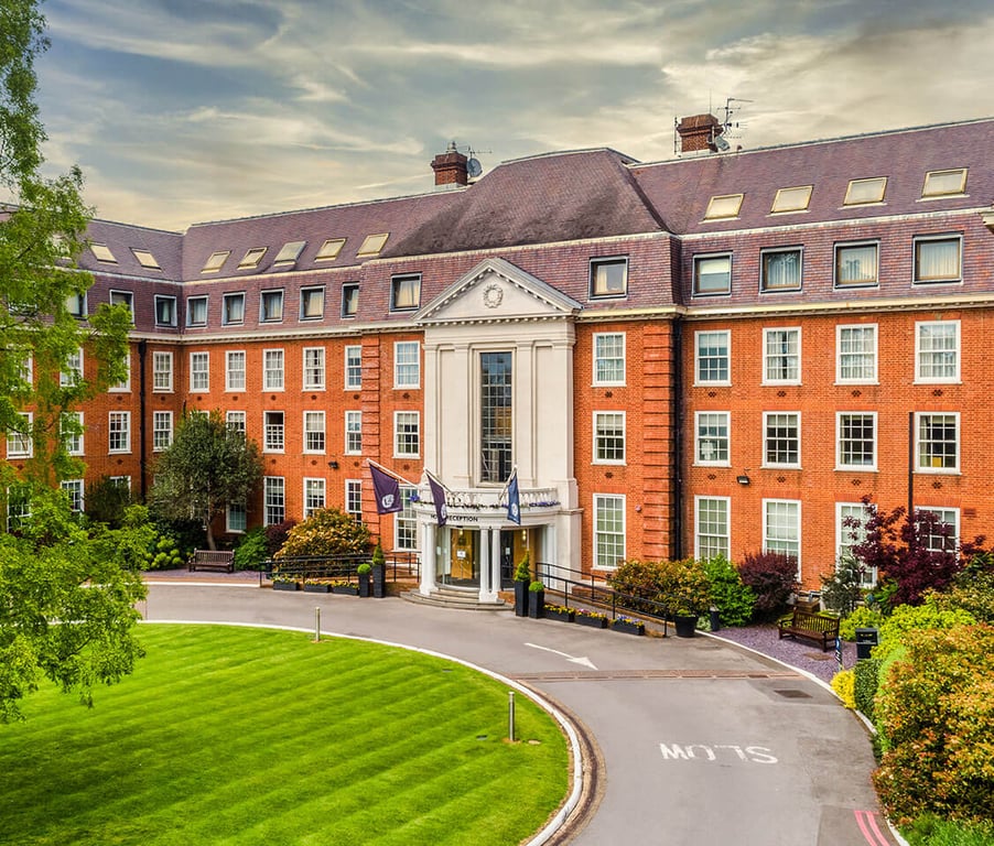 The Lensbury building with a brick facade, columns at the entrance, set amid manicured lawns and greenery under a cloudy sky.