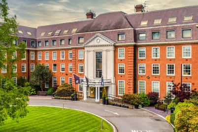 The Lensbury building with a brick facade, columns at the entrance, set amid manicured lawns and greenery under a cloudy sky.