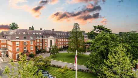 The Lensbury hotel in Teddington, London, with traditional brick architecture, green lawn, and a sunset sky.
