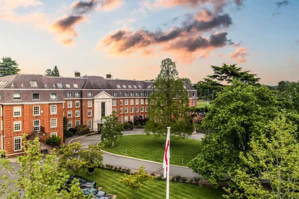 The Lensbury hotel in Teddington, London, with traditional brick architecture, green lawn, and a sunset sky.