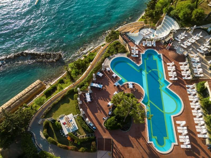 Aerial view of Liberty Lykia Resort's pool area, with a unique pool shape, surrounded by green landscaping and adjacent to the turquoise sea.