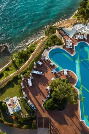 Aerial view of Liberty Lykia Resort's pool area, with a unique pool shape, surrounded by green landscaping and adjacent to the turquoise sea.