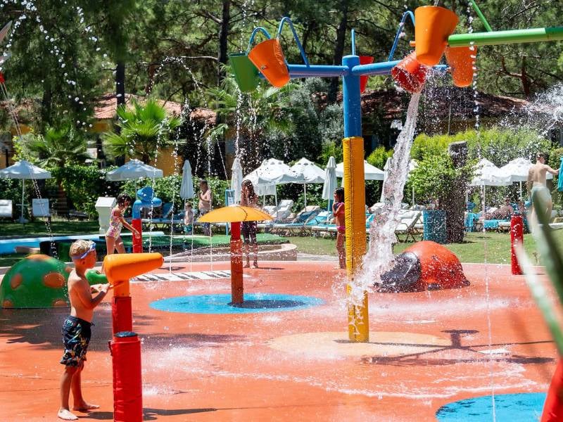 Children playing in the colourful splash park at Liberty Lykia Family Resort, with water jets and buckets against a backdrop of greenery.