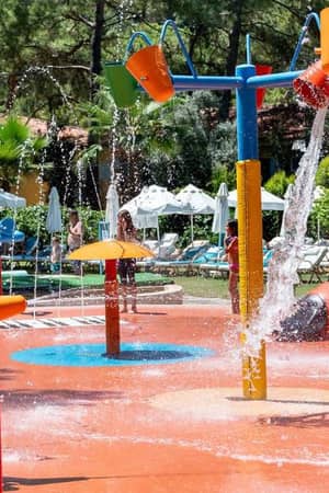 Children playing in the colourful splash park at Liberty Lykia Family Resort, with water jets and buckets against a backdrop of greenery.