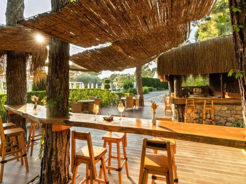 Outdoor bar at Liberty Lykia with wooden stools and bar, surrounded by greenery with woven shaded areas.