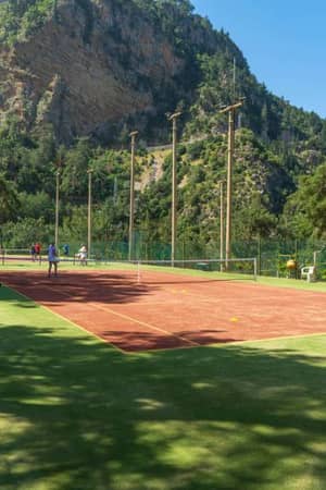 Tennis court at Liberty Lykia Resort with players, set against a backdrop of green mountains and trees, under a clear blue sky.