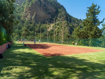 Tennis court at Liberty Lykia Resort with players, set against a backdrop of green mountains and trees, under a clear blue sky.