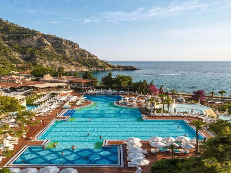 Aerial view of Liberty Lykia's large swimming pool surrounded by sun loungers and umbrellas, with the Aegean Sea and mountains in the background.