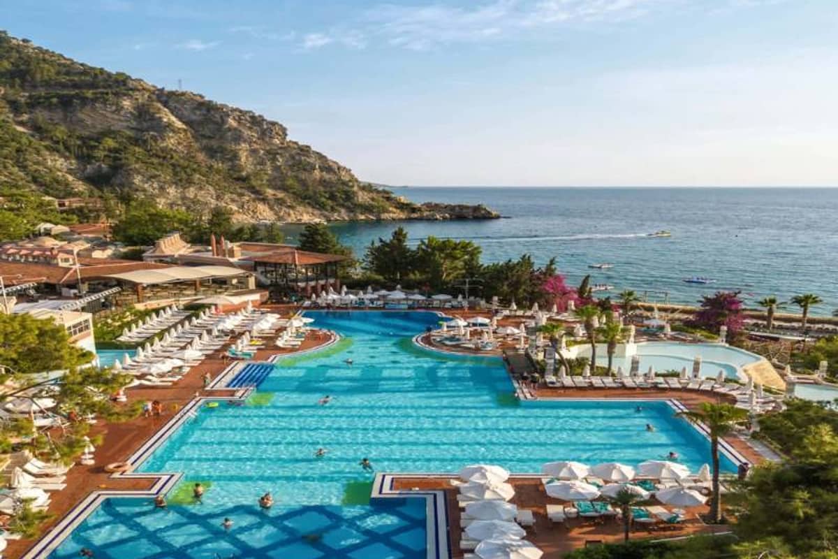 Aerial view of Liberty Lykia's large swimming pool surrounded by sun loungers and umbrellas, with the Aegean Sea and mountains in the background.