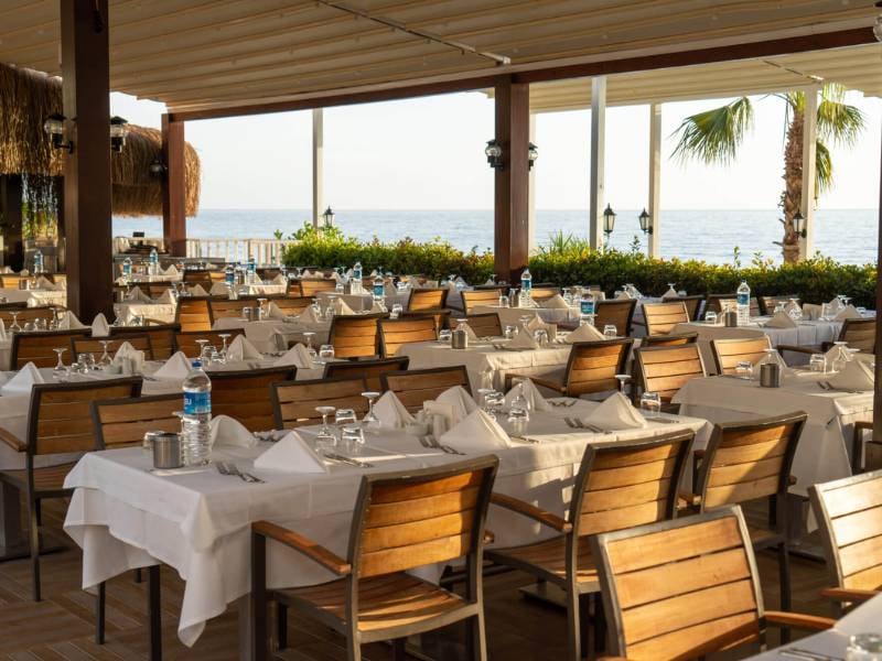 Open-air restaurant at Liberty Lykia Resort with tables set with white tablecloths and wooden chairs, overlooking the sea.