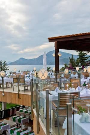 Liberty Lykia rooftop restaurant with sea and mountain views, featuring white tablecloths and wooden seating under a pergola.