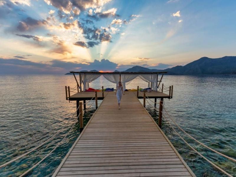 Woman in white atop a wooden pier at sunrise, with sunrays illuminating the sea and mountains at Liberty Lykia.