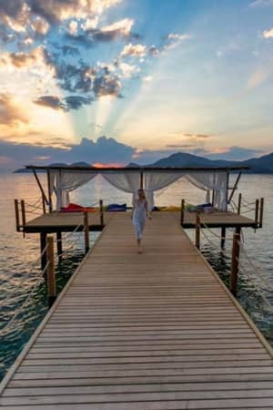 Woman in white atop a wooden pier at sunrise, with sunrays illuminating the sea and mountains at Liberty Lykia.