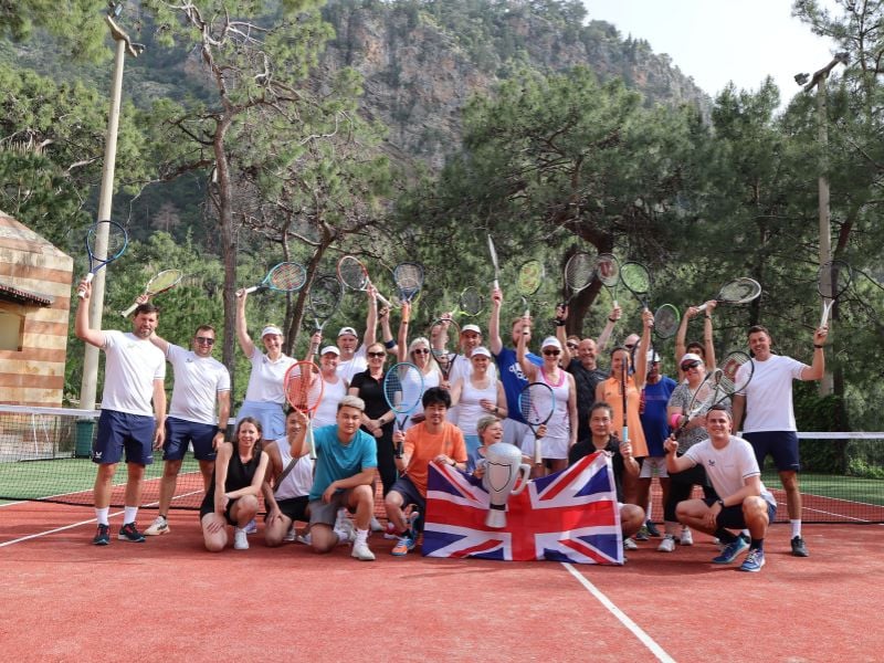 Group of people on a tennis court at Liberty Lykia, holding rackets and a British flag, with trees and mountains in the background.
