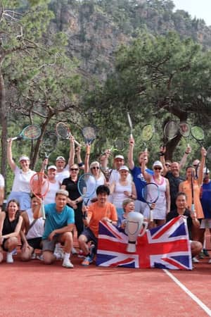 Group of people on a tennis court at Liberty Lykia, holding rackets and a British flag, with trees and mountains in the background.