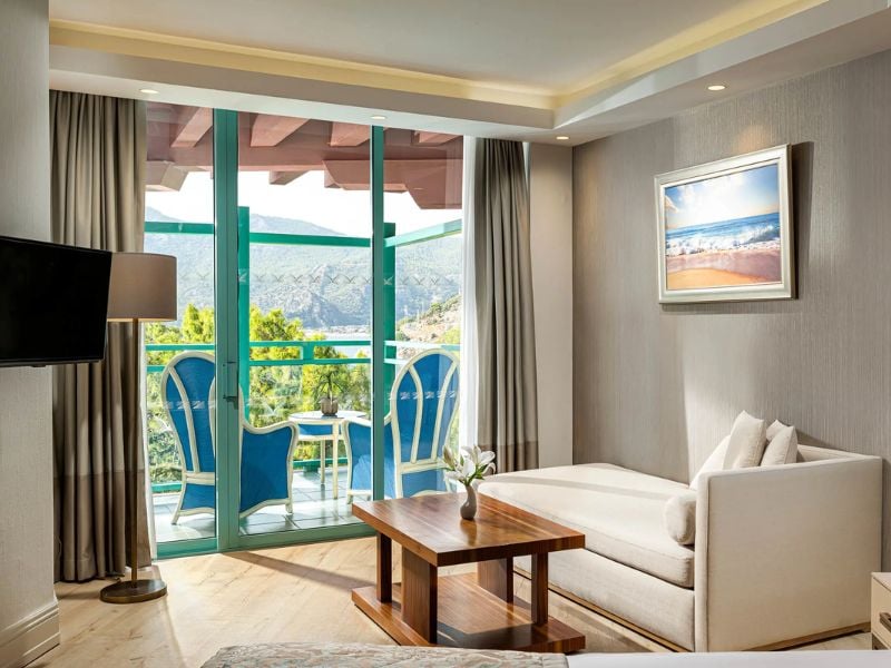 Hotel room at Liberty Lykia Resort with a comfortable lounge area, balcony with blue chairs, and a view of greenery and mountains.
